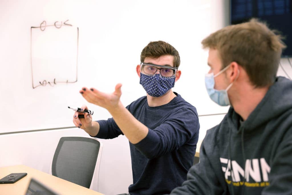 Student gestures with an open hand in front of a whiteboard with a circuit drawn on it