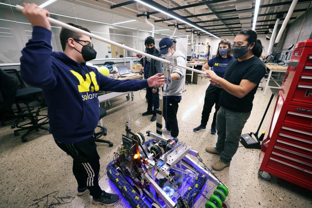 two team members hold either end of a pvc pipe at about shoulder height. Their robot's climbing hook rests over the middle of the pipe