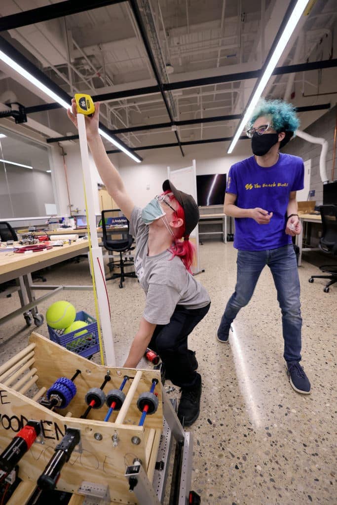 A student crouches next to a boxy robot, extending a tape measure as far upward as she can reach.