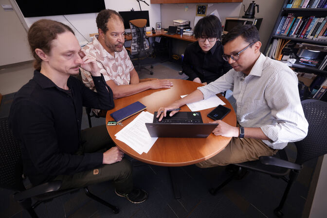 Four men are seated around a small round table, eyes on a laptop screen.