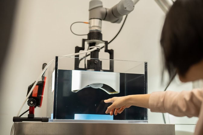 A hand points at a transducer submerged in a water tank in a lab setup.