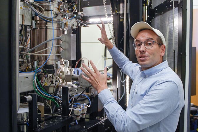 A man gestures toward a complex network of wires, plugs and metal pipes inside a cabinet-like container.