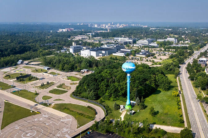 A drone-captured bird’s-eye view. Left foreground: a parking lot and roundabout. Center foreground: Green space and a water tower. Right: a road with two lanes in each direction extends vertically, then curves out of view. Buildings dot the background and line the road on the right.