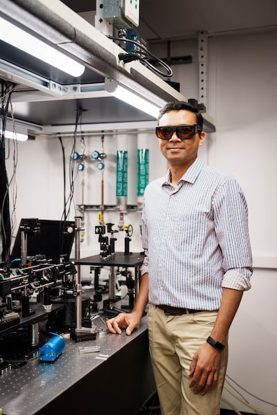 Parag Deotare stands under a set of fluorescent lights, next to an optical table equipped with lasers, detectors and many lenses and mirrors, at different heights. He wears a pair of tinted laser safety glasses.