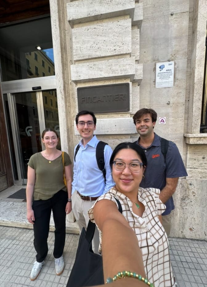 Group of four people taking a selfie outside a building with a "FINCANTIERI" sign.