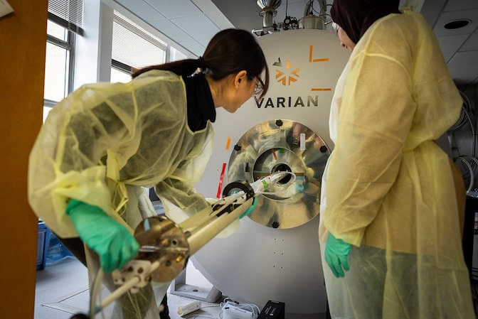 Two people in protective gear work with a large MRI machine labeled "VARIAN" in a lab setting.