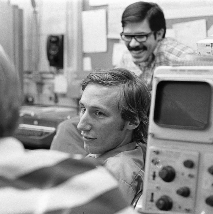 A black and white photo shows two young men smiling in a lab, with an oscilloscope in the foreground.