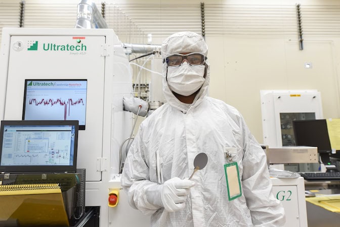 A man wears a PPE bunny suit with a hood, a mask, safety glasses and gloves while standing to the right of a large, refrigerator-sized machine with two computer screens on the surface displaying a line graph. He holds silver tongs in one hand, grasping a thin metallic wafer a few inches in diameter.