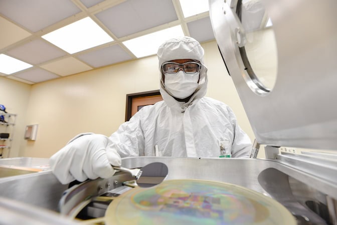 A man wears a PPE bunny suit with a hood, a mask, glasses and gloves while standing in front of an inkjet-printer-sized machine with the top hinged open. One hand holds tweezers and a thin wafer a few centimeters in diameter over a larger, opalescent disc