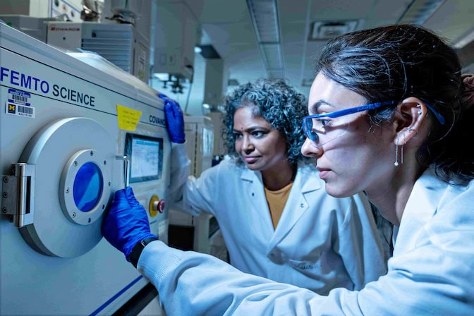  Two women in lab coats read a digital monitor on an oven-like device.