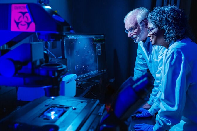 Two scientists view a computer monitor, which displays a series of white cancer cells. The monitor is next to a microscope, which is shining blue-tinted light on a glass slide.