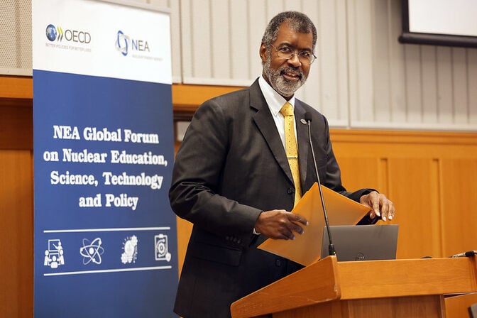 Man speaking at a podium with a conference banner in the background. The banner reads, "OECD NEA Global Forum on Nuclear Education, Science, Technology and Policy."