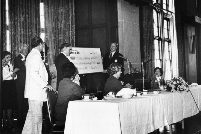 Black and white photo of People presenting and applauding a large ceremonial check at a formal event.