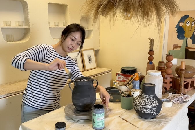  A woman applies black paint to a vase. Another black vase nearby is decorated with white swirls.