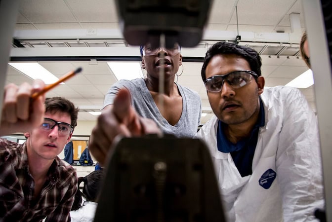 Three people wearing safety glasses concentrate on a laboratory experiment. One person points at the equipment while another holds a pencil, all focusing intently on the setup.