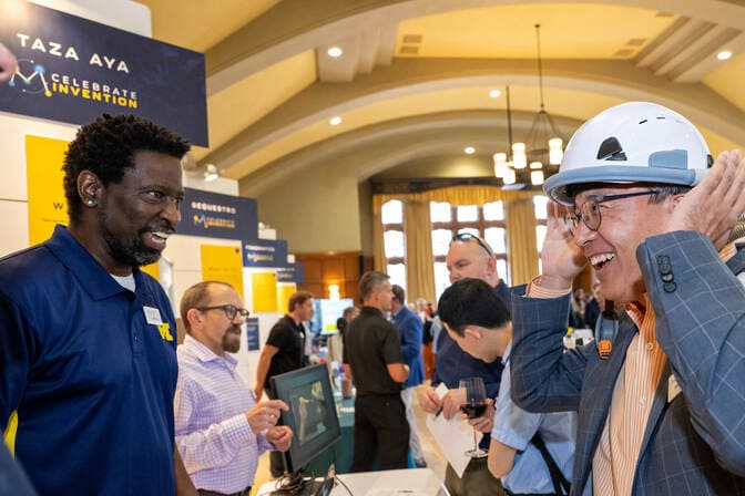 Two men interacting at a conference; one wears a hard hat and the other a navy polo shirt.