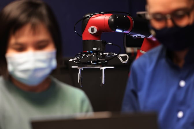 Foreground: Two out of focus researchers look at a computer screen together. Background: An in focus, red robotic arm stands on a pedestal.