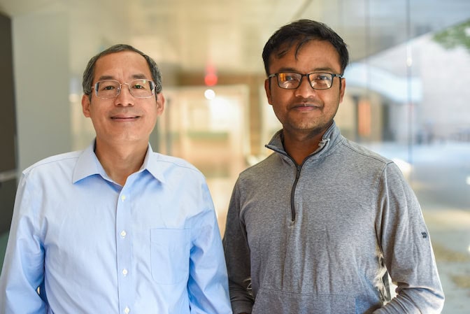 Portrait of two men standing side by side in a long, white hallway with floor-to-ceiling glass windows on one side.