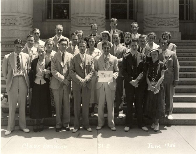 A black and white group photo from 1936 of people in formal attire standing on steps in front of a neoclassical building. One man in the front center is holding a paper with “31E” written on it.