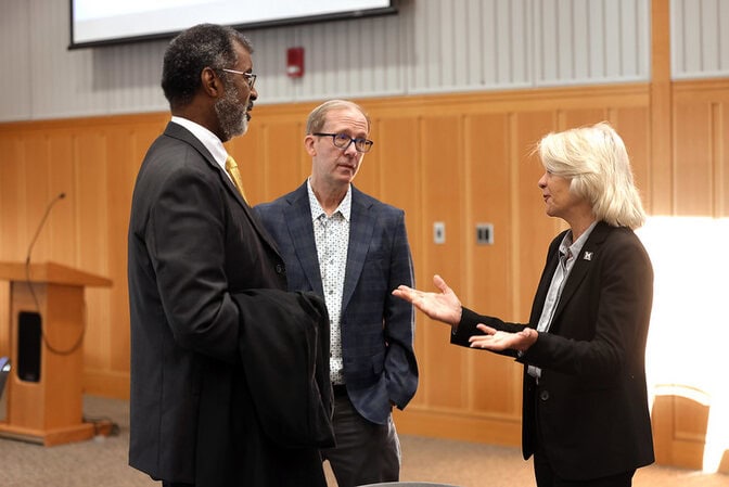 Two men and a woman, standing, converse in a conference room.