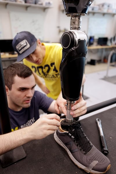 Seelhoff appears to adjust the athletic shoe covering the foot of the prosthesis. Best leans in to steady the prosthesis with a hand on the metal post that links the foot to the black, calf-shaped box housing the motor and electronics.