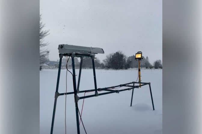 An untouched, snowy field with trees in the background. In the foreground, a long camera is set up on tall, metal poles. A light stands on a pole about two meters in front of the camera.