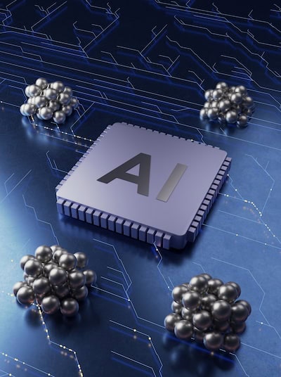 A gray computer chip with the letters “AI” on top in black sits on a circuit board with neural network lines and nodes etched in white. Four clumps of small silver spheres sit on the metal surface surrounding the chip.