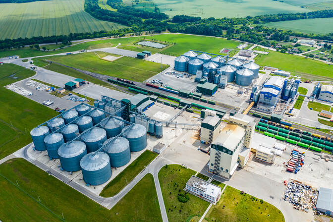 An aerial view of a bioethanol facility. Green fields surround a facility with silver drums several stories high positioned in grids. Two tall buildings sit to the right of the silver drums. A road divides two sets of silver drums with tractor trailers ready to transport biofuel.
