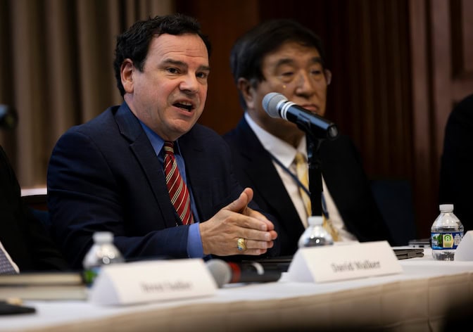 A man sitting at a long table speaks into a microphone.