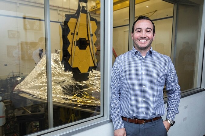 Robby Swoish standing by a window with a scientific setup involving hexagonal gold plates behind him.