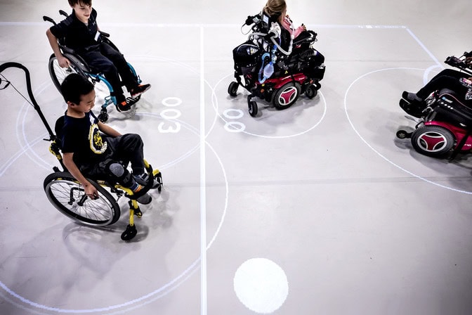 Two children in manual wheelchairs play against peers in power chairs, the ball appearing as a white spot in an open space. The lines from the projector bound a rectangle around them and the center line between them as well as arcs marking the goals.