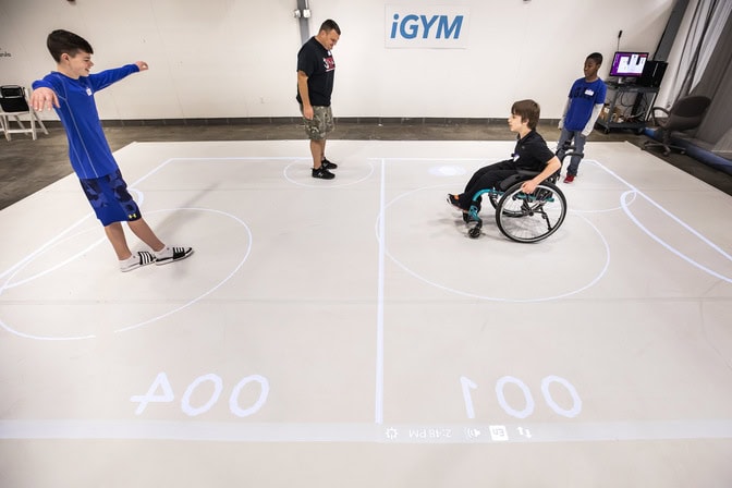 At an iGYM game demo at U-M, a child in a wheelchair faces off with another who can walk. A projector shows the location of the digital ball and the boundaries of the field. The circles around the players expand to represent kicks. Credit: Joseph Xu/Michigan Engineering