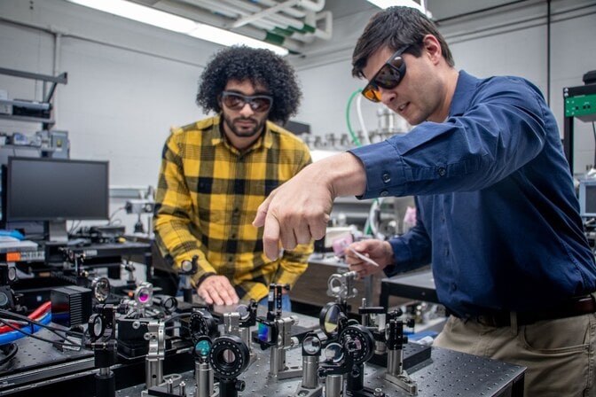 Two men in dark protective glasses work at an optical table, a black surface with optical components like lenses and reflectors secured to it. 