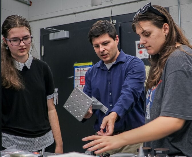 Limbach gestures toward the table while Carman reaches for something out of frame. Watts observes.