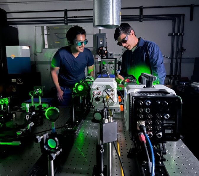 Two men in dark protective glasses stand at a blocky white machine with cables extending from the back. The optical components on the table glow green with laser light.