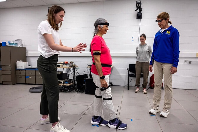 Center: A woman stands on one leg while wearing black velcro straps around her head, upper arms, wrists, hips, upper thighs, below the knees and feet. Left: A woman stands behind with her hands stretched to catch if she falls. Right: Two women observe.