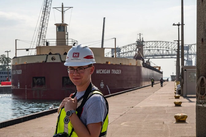A man wearing a hard hat, neon yellow safety vest, stands in front of a large barge at a dock. A DSLR camera is strapped around the man's neck.