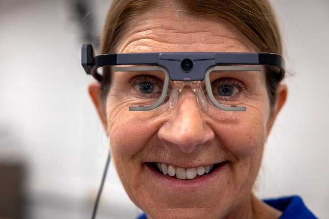 A close up of a smiling woman wearing glasses with a solid plastic bar across the forehead and a camera in the center of the bar. Gray, half rectangles with no lens surround the eyes where the lens would be in eyeglasses.