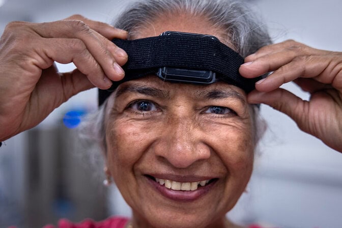 A closeup of a smiling woman’s face. She uses her hands to adjust a black velcro strap positioned around her forehead. The strap has a square, black plastic device in the center.