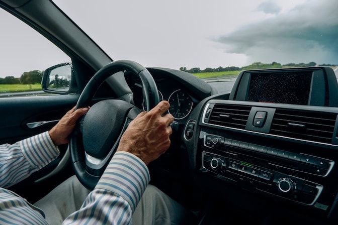 Hands with prominent veins on a steering wheel in the foreground. Outside the vehicle, heavy clouds are rolling in.