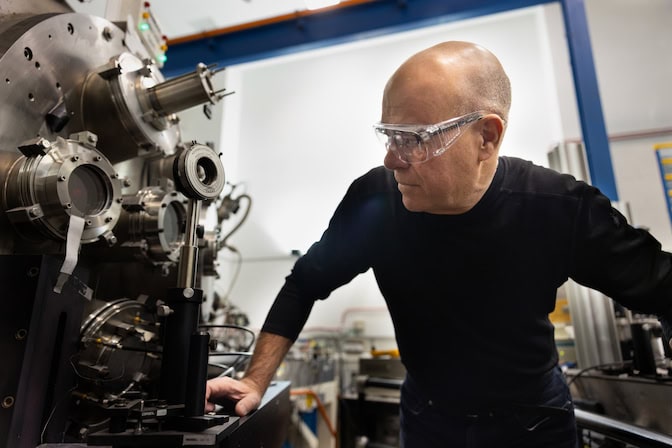 A man, wearing safety goggles, peers at a lens mounted on a metal rod. A large metal dome with several, round glass portholes is behind the lens.