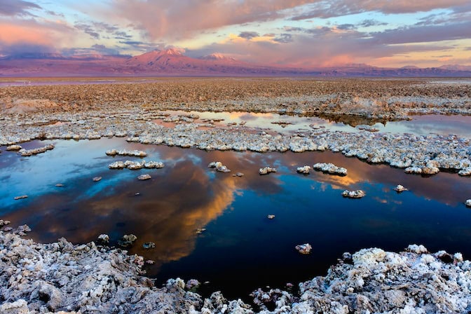 A shallow lake holds the reflections of clouds, highlighted by a pink and orange sunset. White-gray rocks protrude from the water's surface in islands and ridges.