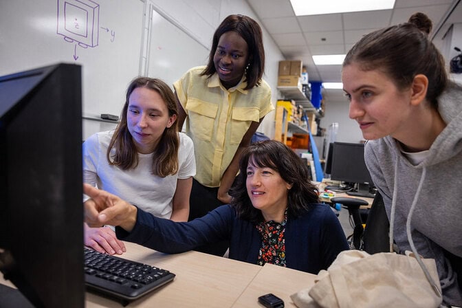 Four women gather together to look at a computer monitor in the foreground. One woman points and speaks while the others look on.