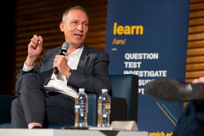 Thomas Zurbuchen sits on stage, holding a microphone while speaking to an audience and discussing with another person. There is blue banner with the word, “learn,” displayed on it in the background.