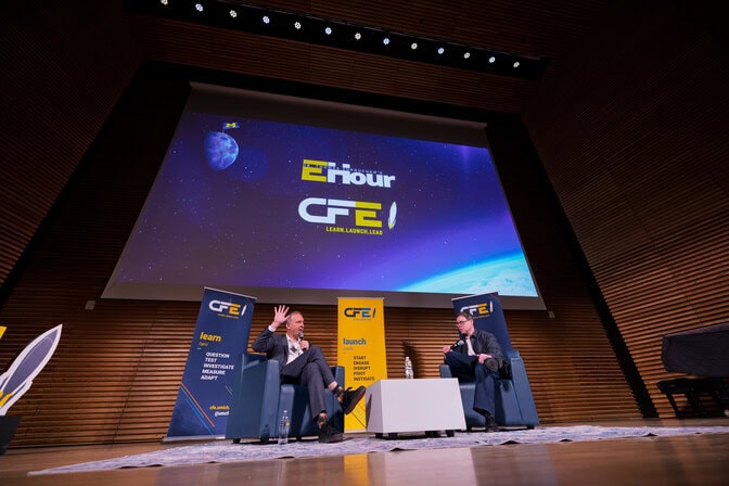 Two individuals seated on stage at a University of Michigan event with a presentation projection and CFE banners behind them.