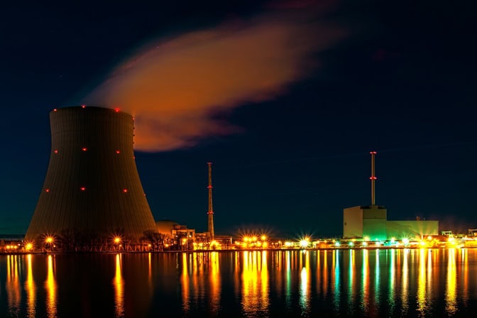 A nighttime view of a power plant with a cooling tower, emitting vapor, reflecting on nearby water.
