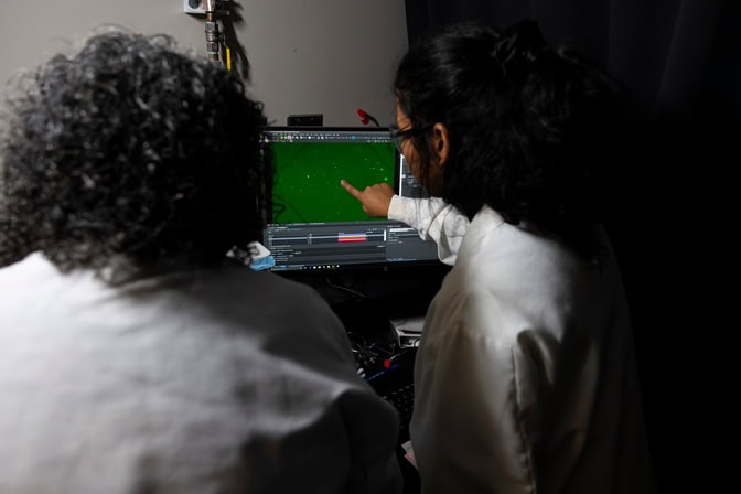 Kumari and Nagrath are seen from the back, with Kumari pointing at a green screen speckled with small, bright dots, zoomed in on a single microwell.