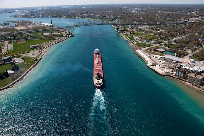 An aerial view of a large freighter cruising down a dark blue river. Each side of the river is flanked by urban areas.