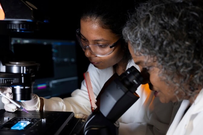 Kumari rests a hand on the microscope, with the chip illuminated nearby. Nagrath looks into an eyepiece similar in size and shape to binoculars.