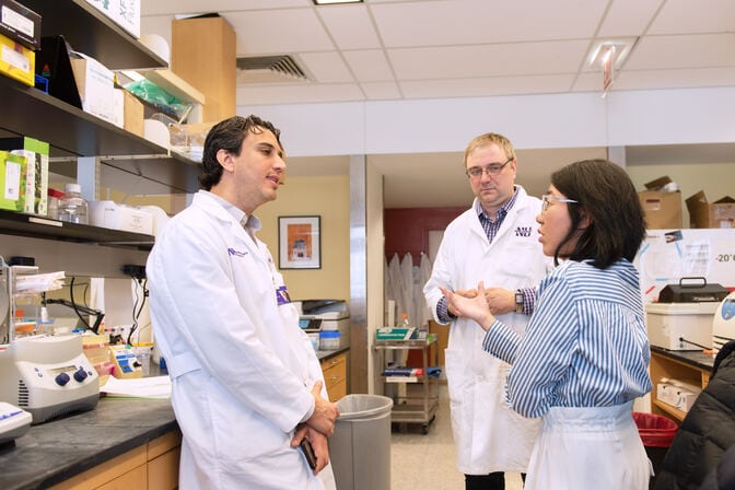 Sonabend leans against a lab bench, listening to a colleague while another looks on.